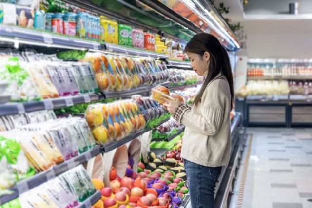 A woman examines packaged fruit in a grocery store aisle filled with various produce.