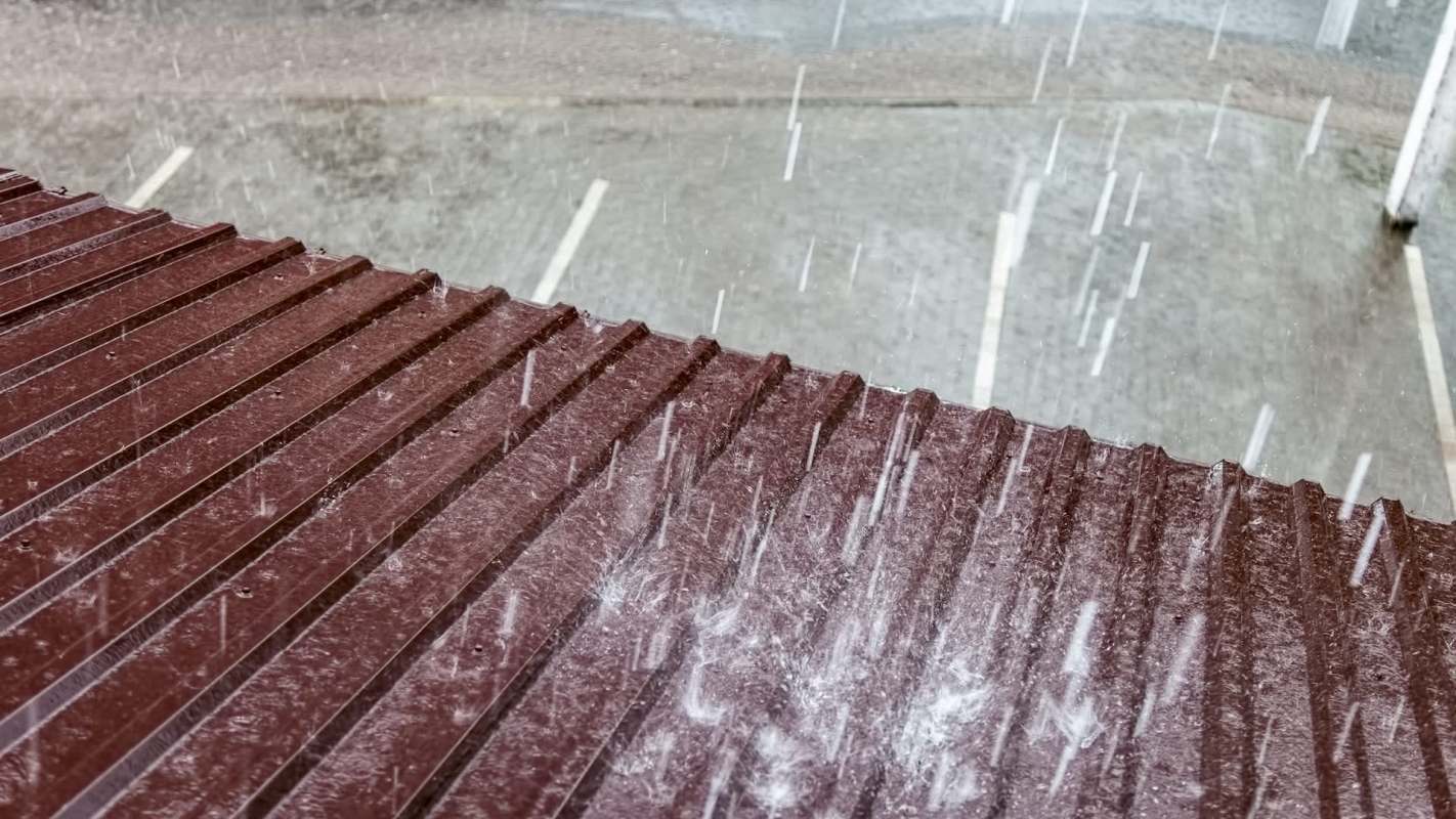 Rain falls on a sloped metal roof, with a blurred background of a wet pavement and parking spaces.