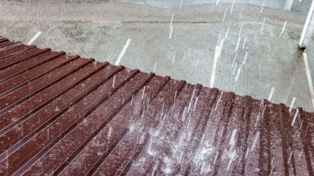 Rain falls on a sloped metal roof, with a blurred background of a wet pavement and parking spaces.