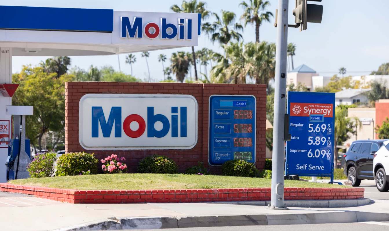 A Mobil gas station sign displaying fuel prices, with $5.59 and $5.69 as the cheapest prices, and surrounded by greenery.