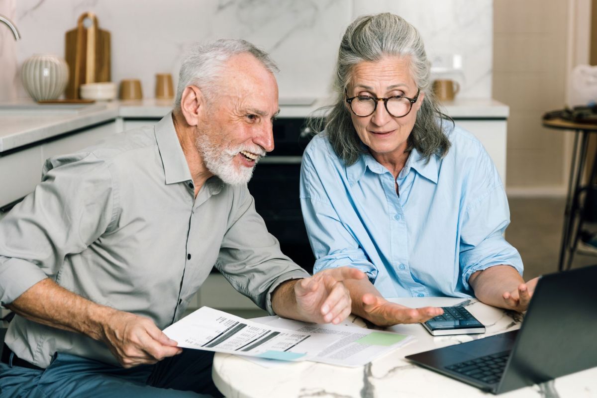 An older couple discusses documents while sitting at a kitchen table with a laptop and calculator.