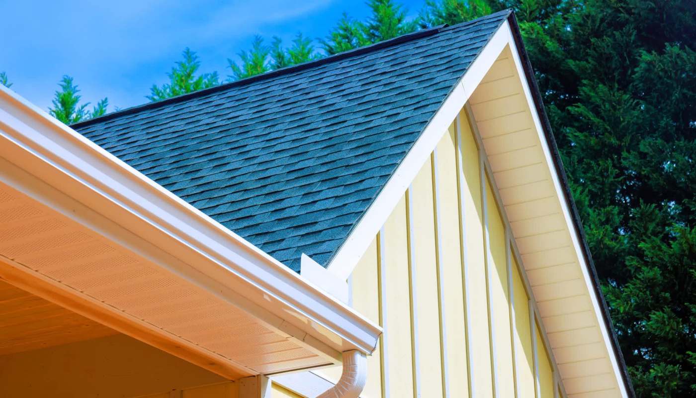 A corner of a yellow house featuring a black shingle roof and a white gutter under a blue sky.