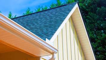 A corner of a yellow house featuring a black shingle roof and a white gutter under a blue sky.