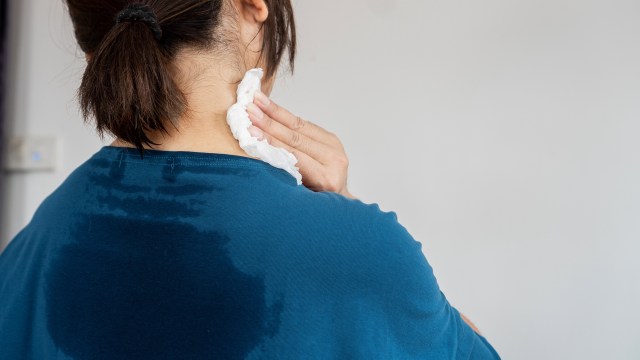 A person wipes their neck with a tissue while wearing a damp blue shirt.