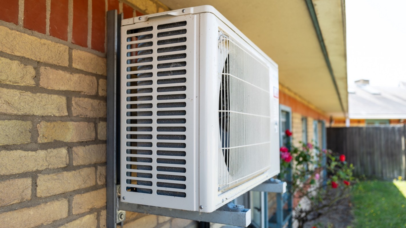 An air conditioning unit mounted on a brick wall next to a blooming rose bush.