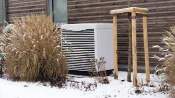 An outdoor area with snow, a large HVAC unit, ornamental grasses, and a wooden tree support structure.