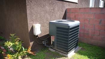 An outdoor air conditioning unit next to a wall, with some greenery in the foreground.