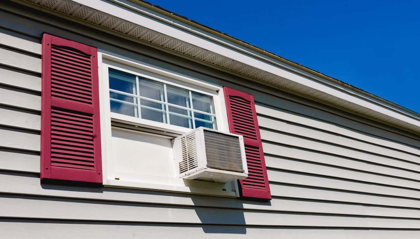 An air conditioning unit installed in a window with red shutters on a house.