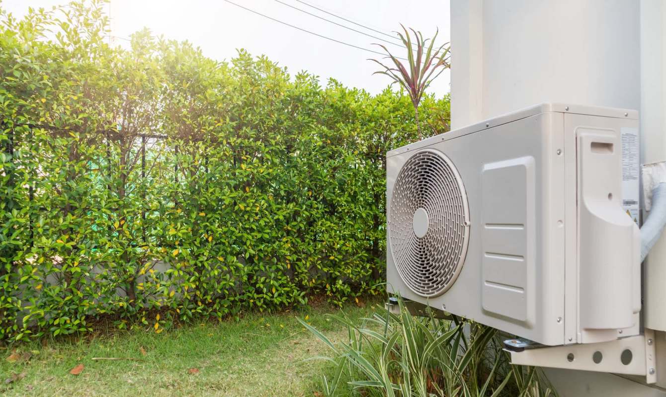 A heat pump unit is installed beside a lush green hedge, with bright sunlight in the upper left corner.