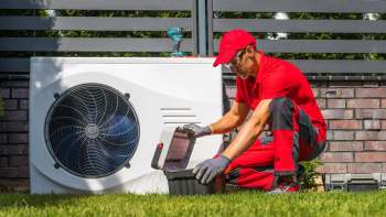 A technician inspects a heat pump unit while using a toolbox in a grassy area.