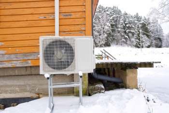 An heat pump unit mounted on a stand next to a wooden building covered in snow.