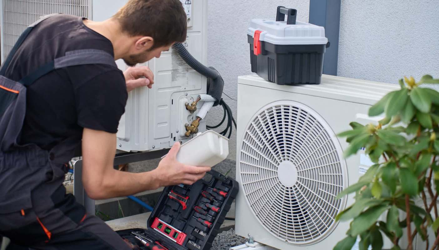 A technician works on an outdoor air conditioning unit with tools nearby.