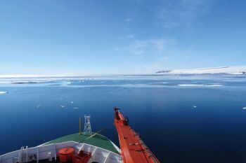 A view from a ship's bow over a calm ocean surrounded by icebergs and snow-capped land under a clear blue sky.