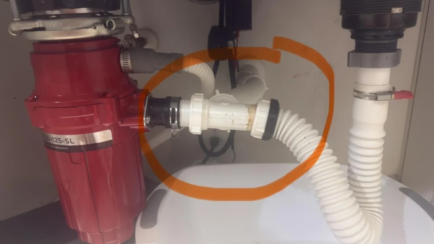 A red garbage disposal unit connected to white and gray pipes under a sink.