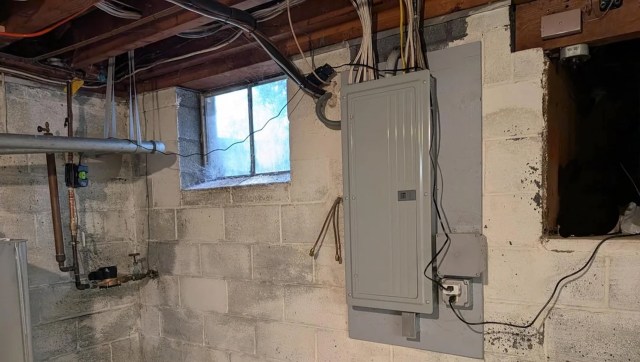 A basement with exposed electrical wiring, a window, and a grey electrical panel on the wall.