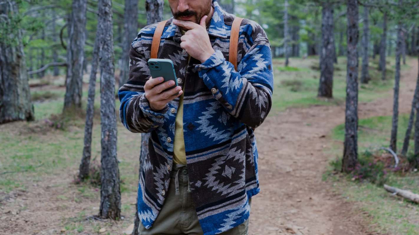 A person checks their smartphone while standing on a forest path among trees, with their hand on their chin.