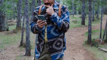 A person checks their smartphone while standing on a forest path among trees, with their hand on their chin.