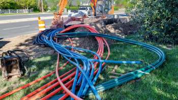 Colored fiberoptic cables are coiled on the ground near construction equipment and a traffic cone, beside a road.