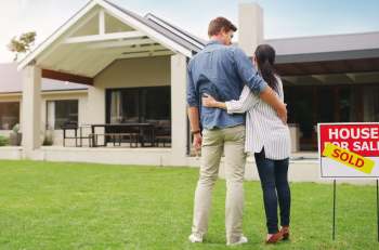 A couple stands in front of a house with a "SOLD" sign in the yard.