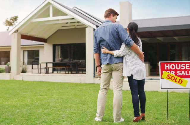 A couple stands in front of a house with a "SOLD" sign in the yard.