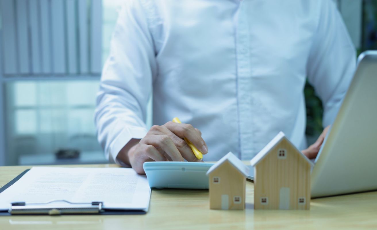 A man sits at a laptop and uses a calculator next to two small wooden houses
