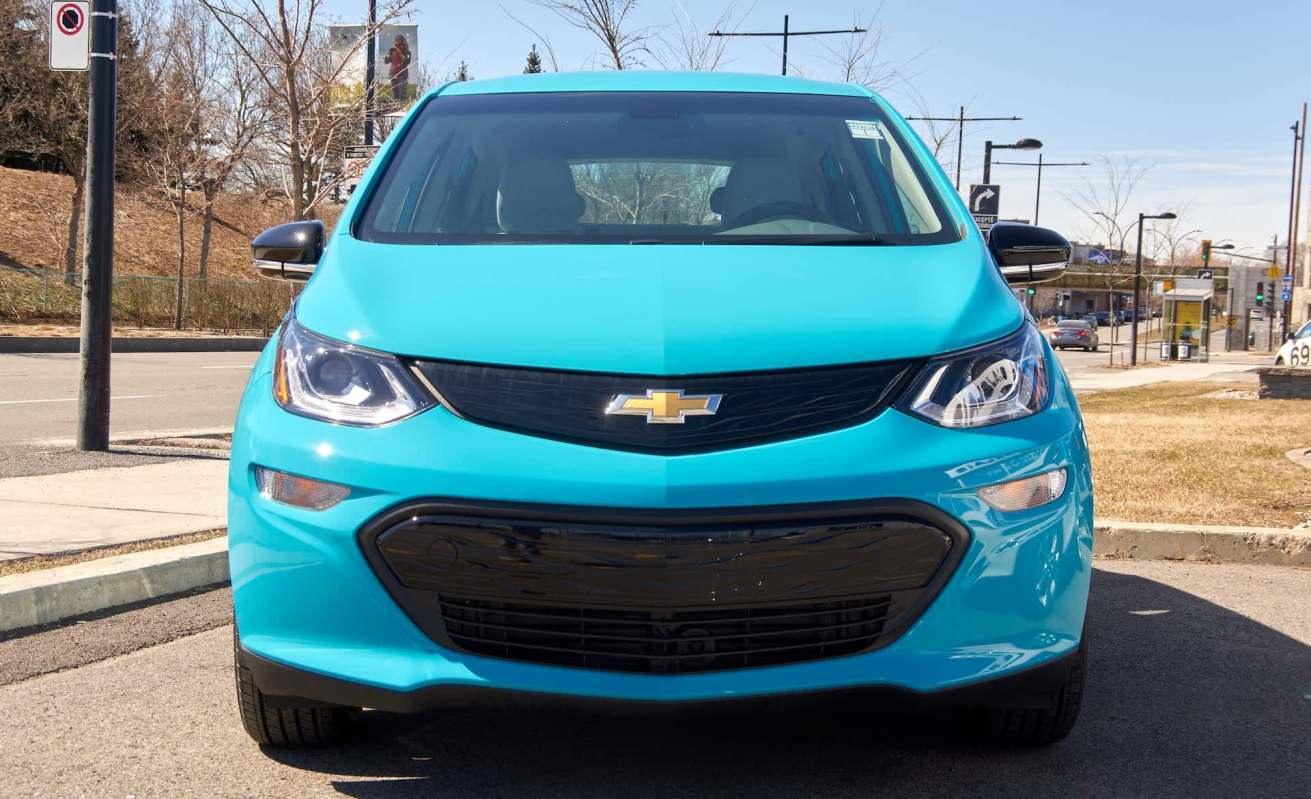 A turquoise Chevrolet Bolt electric car parked on a street with trees and a billboard in the background.