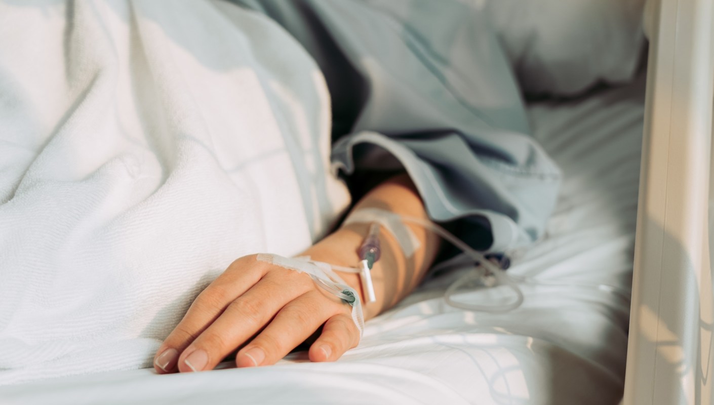 A hand with an IV is resting on a hospital bed covered with a white blanket.