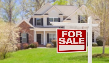 A "For Sale" sign in front of a house on a green lawn.