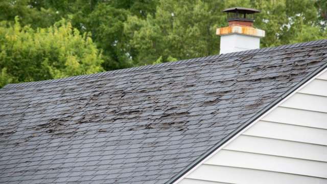 A roof with damaged and curling shingles, alongside a chimney, surrounded by green trees.