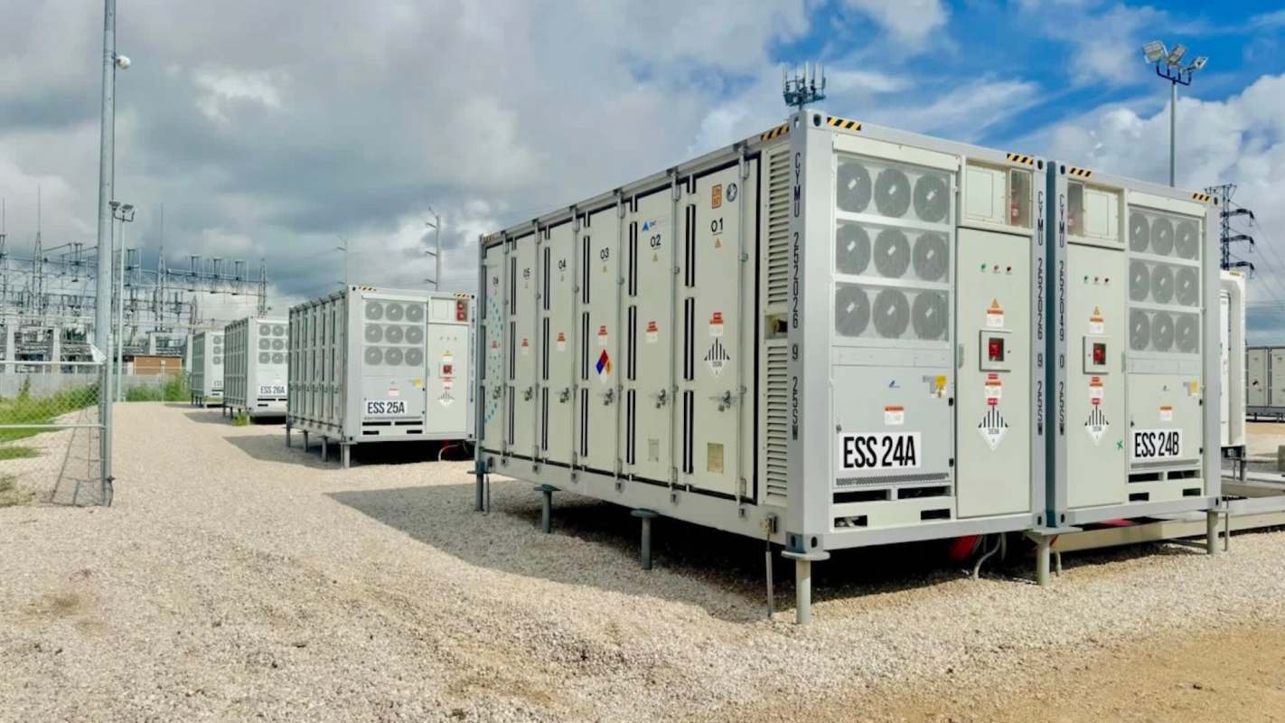 Several battery energy storage containers lined up in a gravel area near a power station.
