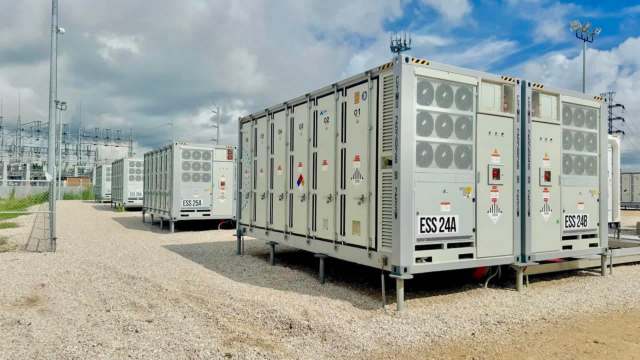 Several battery energy storage containers lined up in a gravel area near a power station.