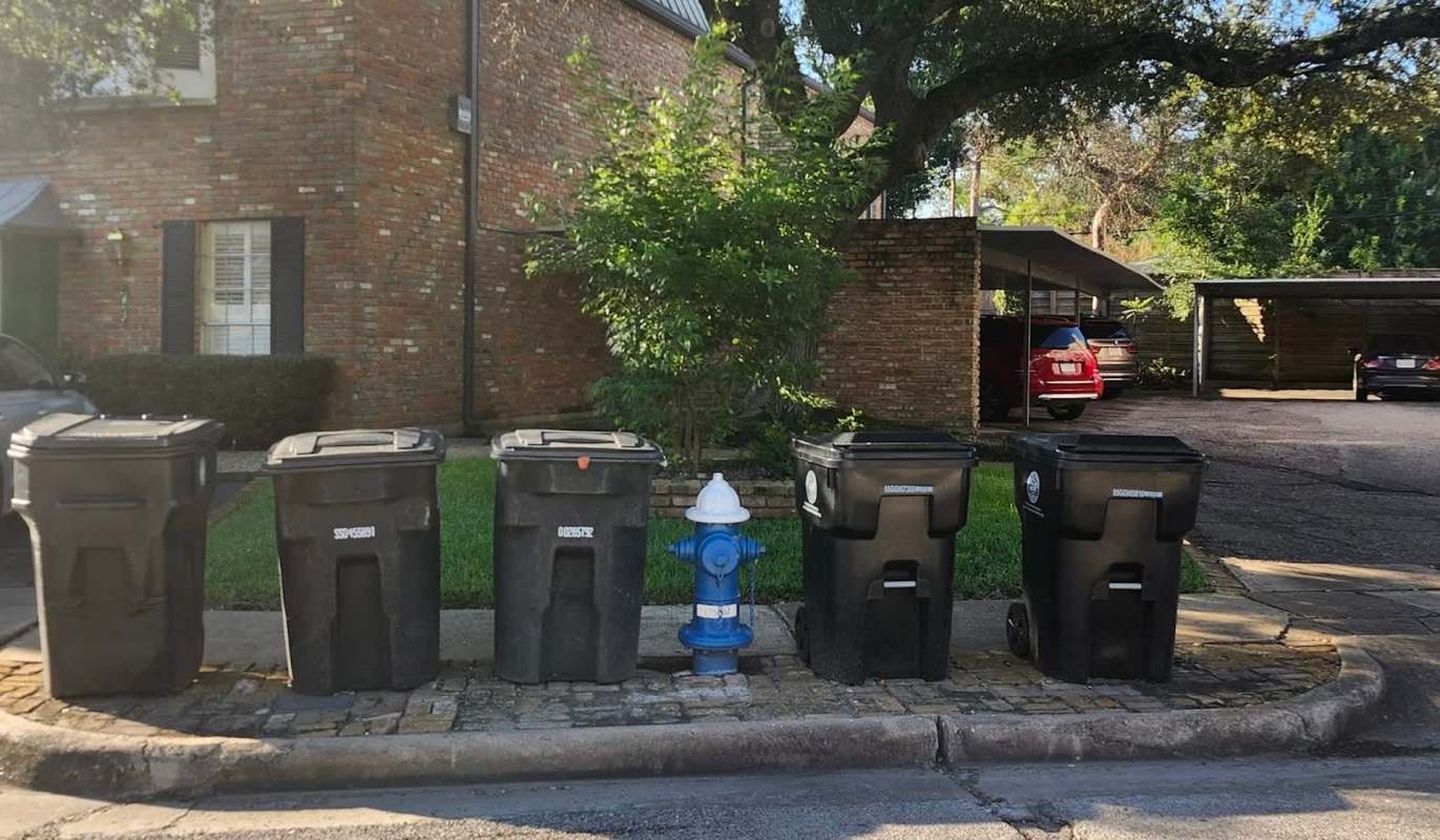 Five trash bins lined up next to a blue fire hydrant in front of a brick building.