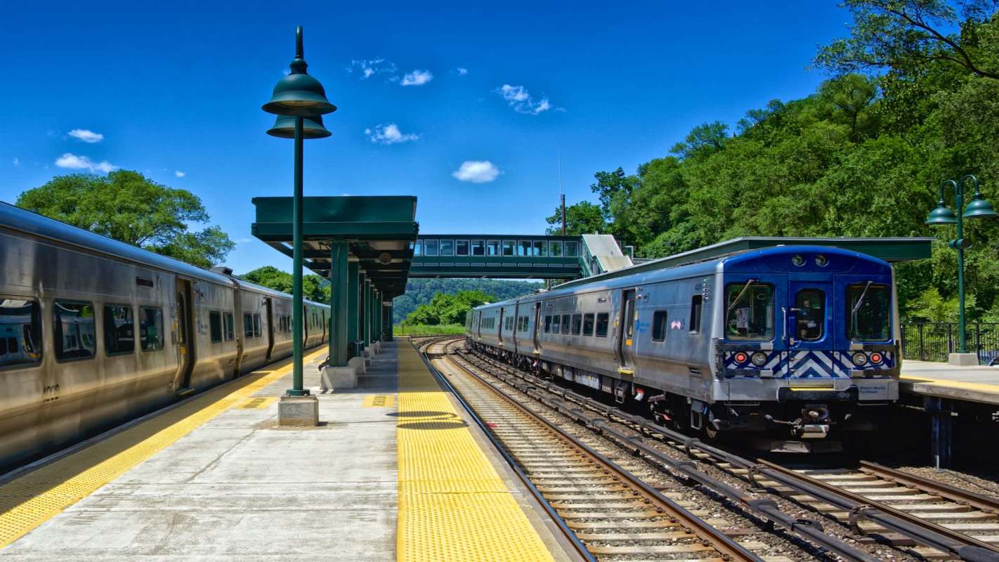 A train arrives at a station with a green overpass and a clear blue sky in the background.