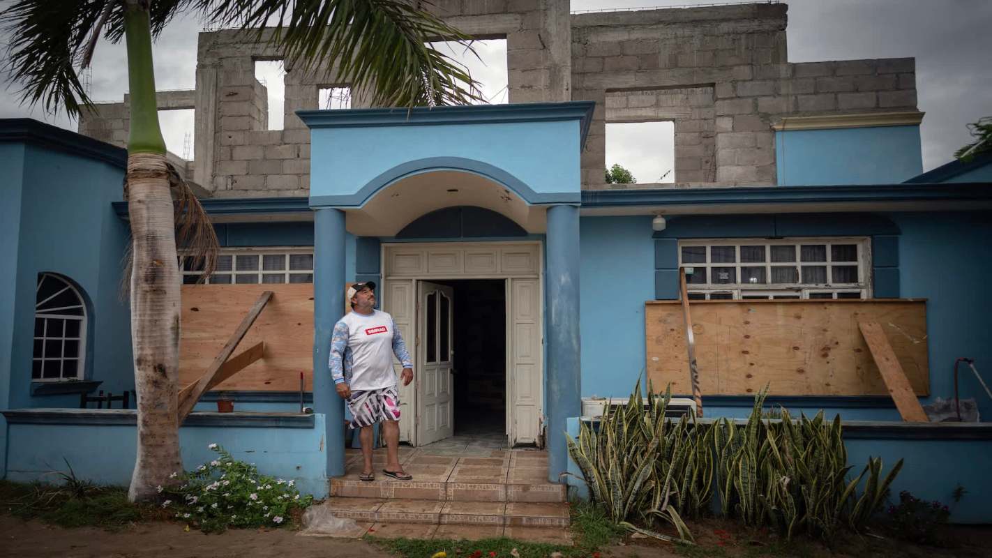 A man standing outside a blue house with boarded windows.