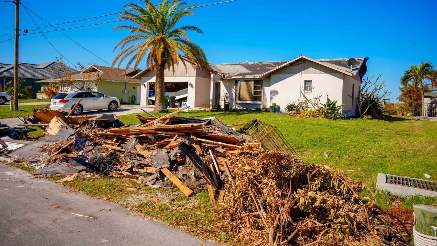 A pile of debris and a palm tree in front of a house with a damaged roof.