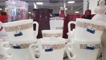 Several white cups with floral designs and price tags are stacked among various kitchen items in a thrift store.