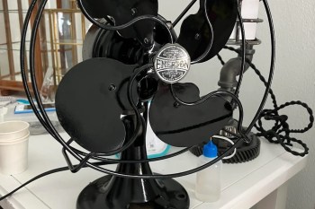 A vintage black Emerson table fan with a circular base and metal blades, displayed on a table with other objects.