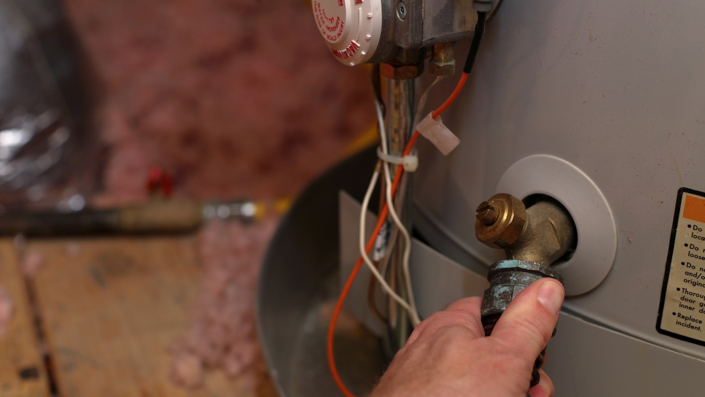 A hand operates a valve on a water heater in a close setting with insulation and pipes visible.