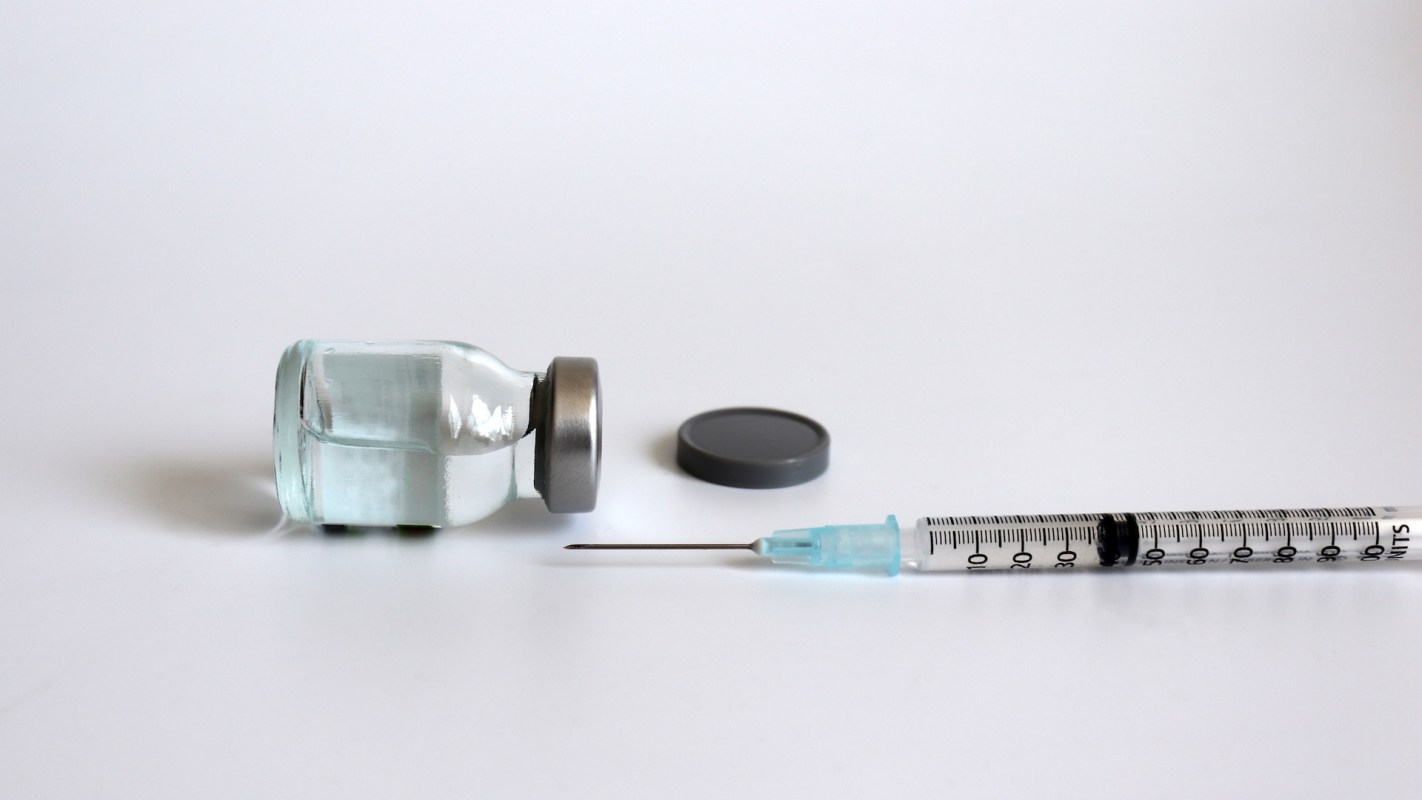 A glass vial with a metal cap beside a syringe on a white surface.