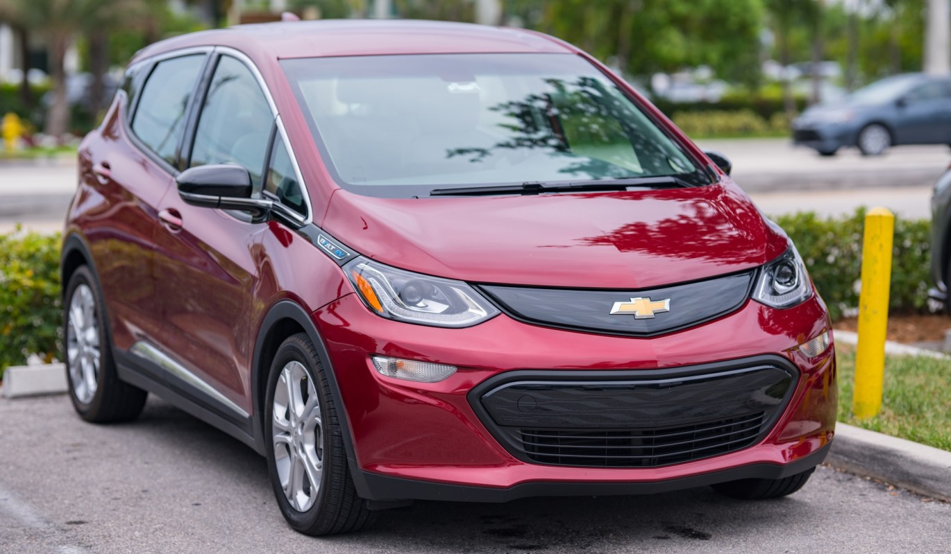 A red Chevrolet Bolt EV parked in a lot with greenery in the background.