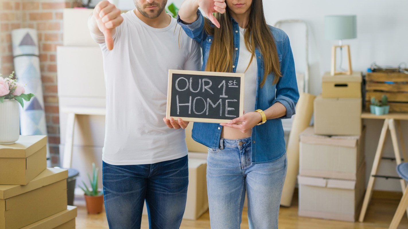 A man and a woman stand in a room holding a chalkboard sign that says "OUR 1st HOME," both giving thumbs down.