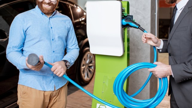 Two men handle a charging cable at an electric vehicle charging station inside a commercial space.