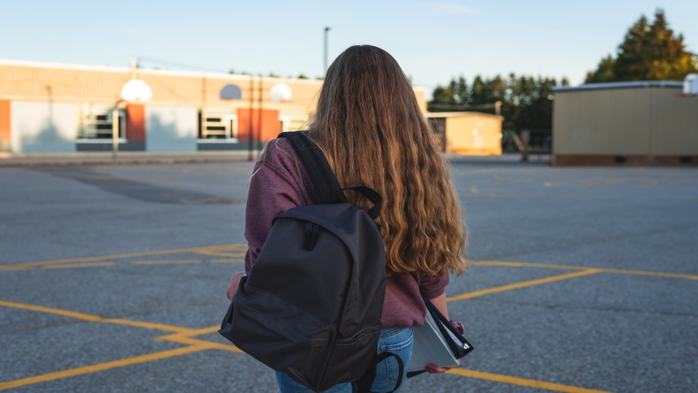 A person walks away from a school building while carrying a backpack and folders.