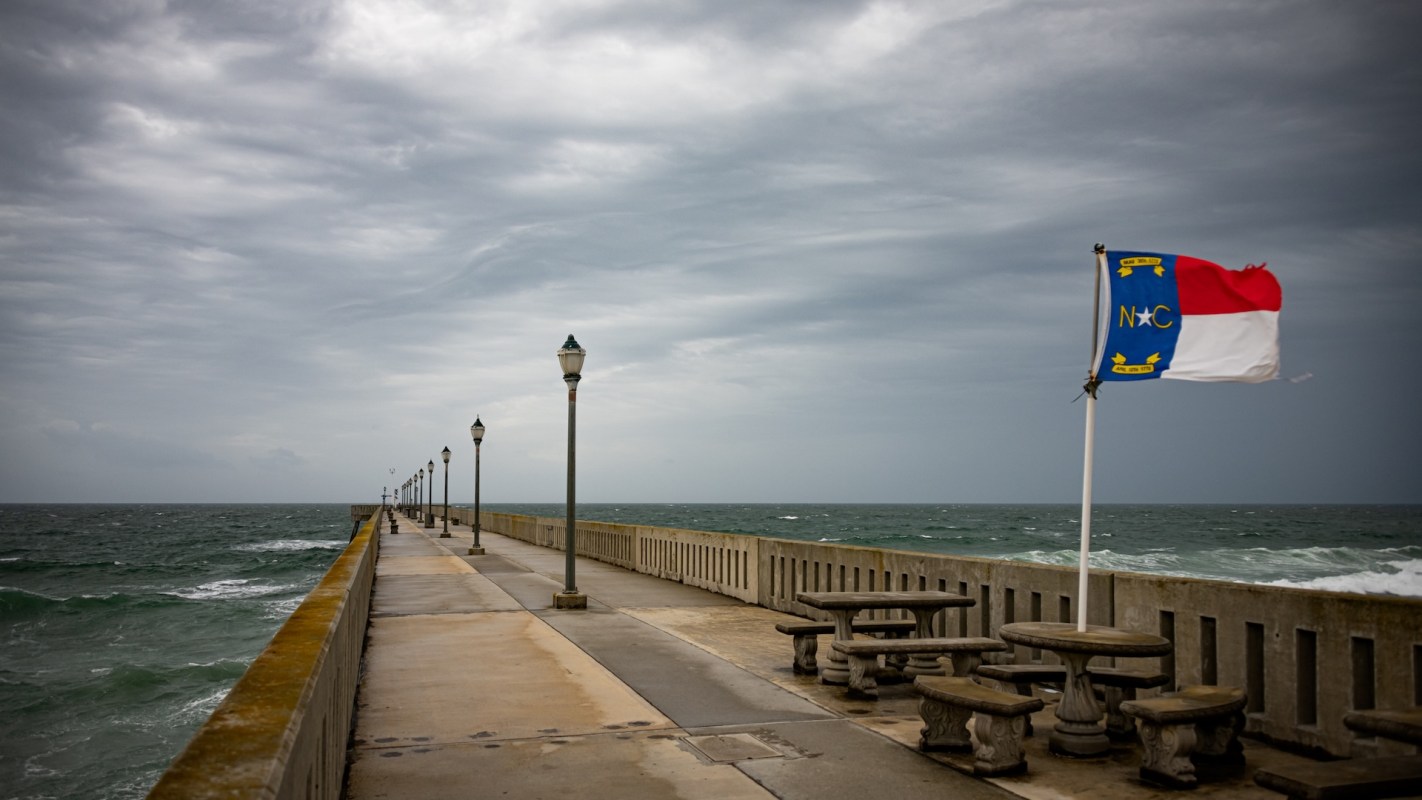 A pier extending over the ocean, lined with lamps and featuring a North Carolina flag, under a cloudy sky.