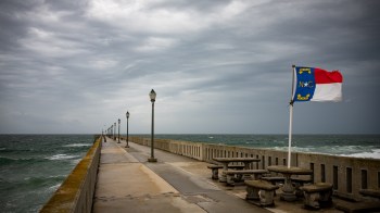 A pier extending over the ocean, lined with lamps and featuring a North Carolina flag, under a cloudy sky.