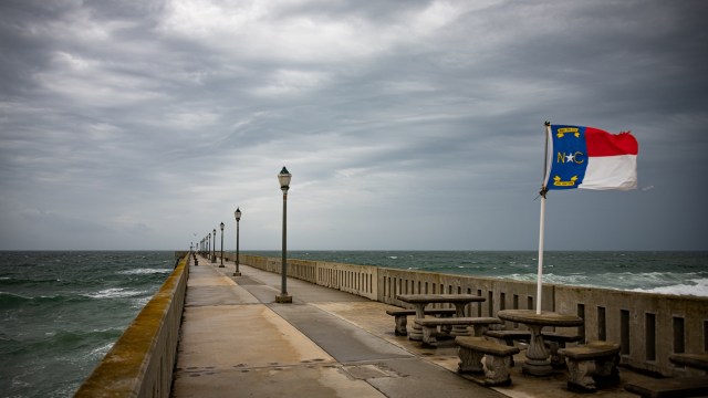 A pier extending over the ocean, lined with lamps and featuring a North Carolina flag, under a cloudy sky.