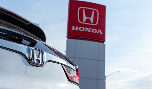 A Honda car with the logo visible in front of a Honda dealership sign.