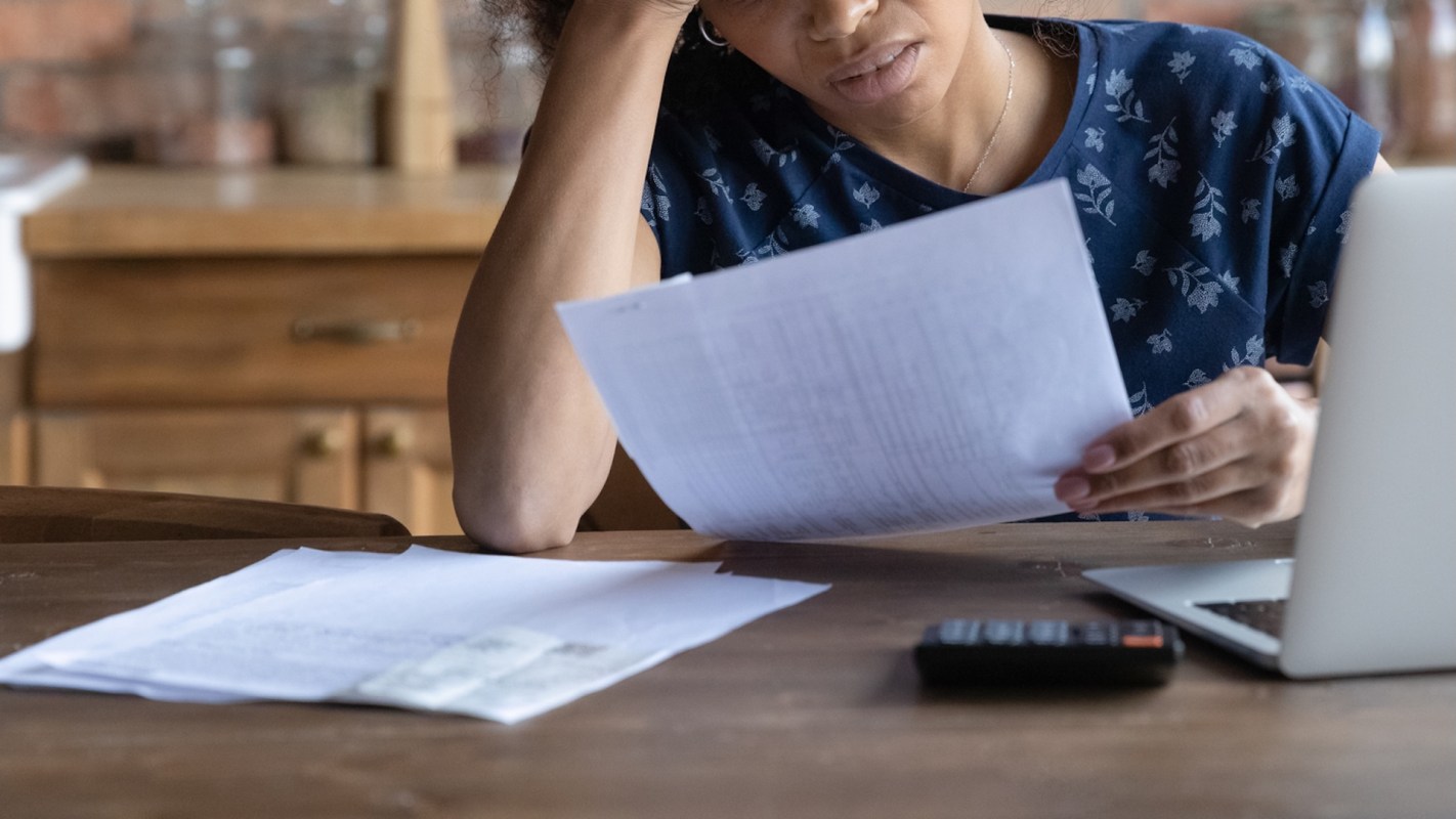 A person, with their hand on their head, reviews documents at a wooden table with a laptop and a calculator nearby.