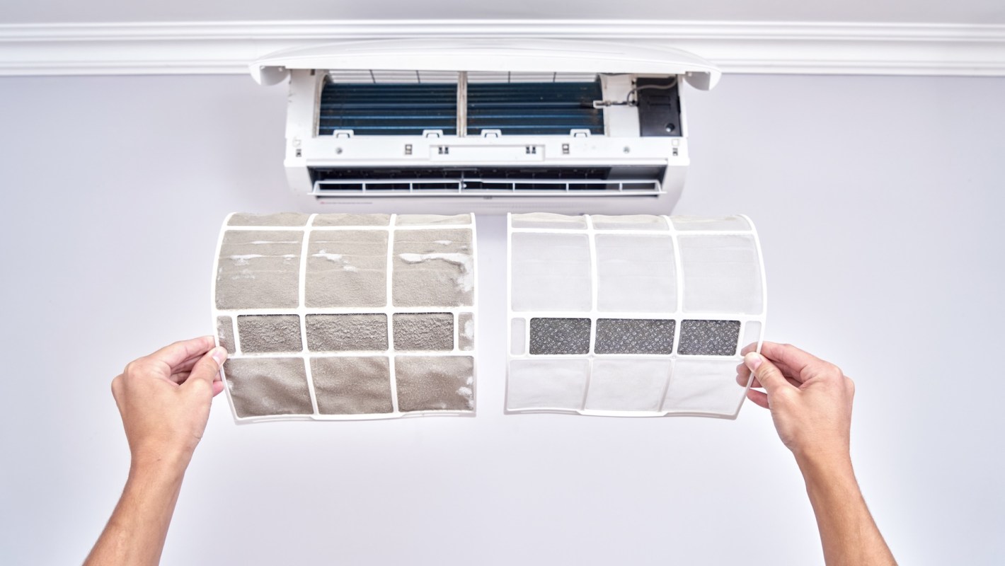 Two air conditioning filters are held up, one dirty and one clean, with an air conditioning unit mounted above.