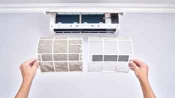 Two air conditioning filters are held up, one dirty and one clean, with an air conditioning unit mounted above.